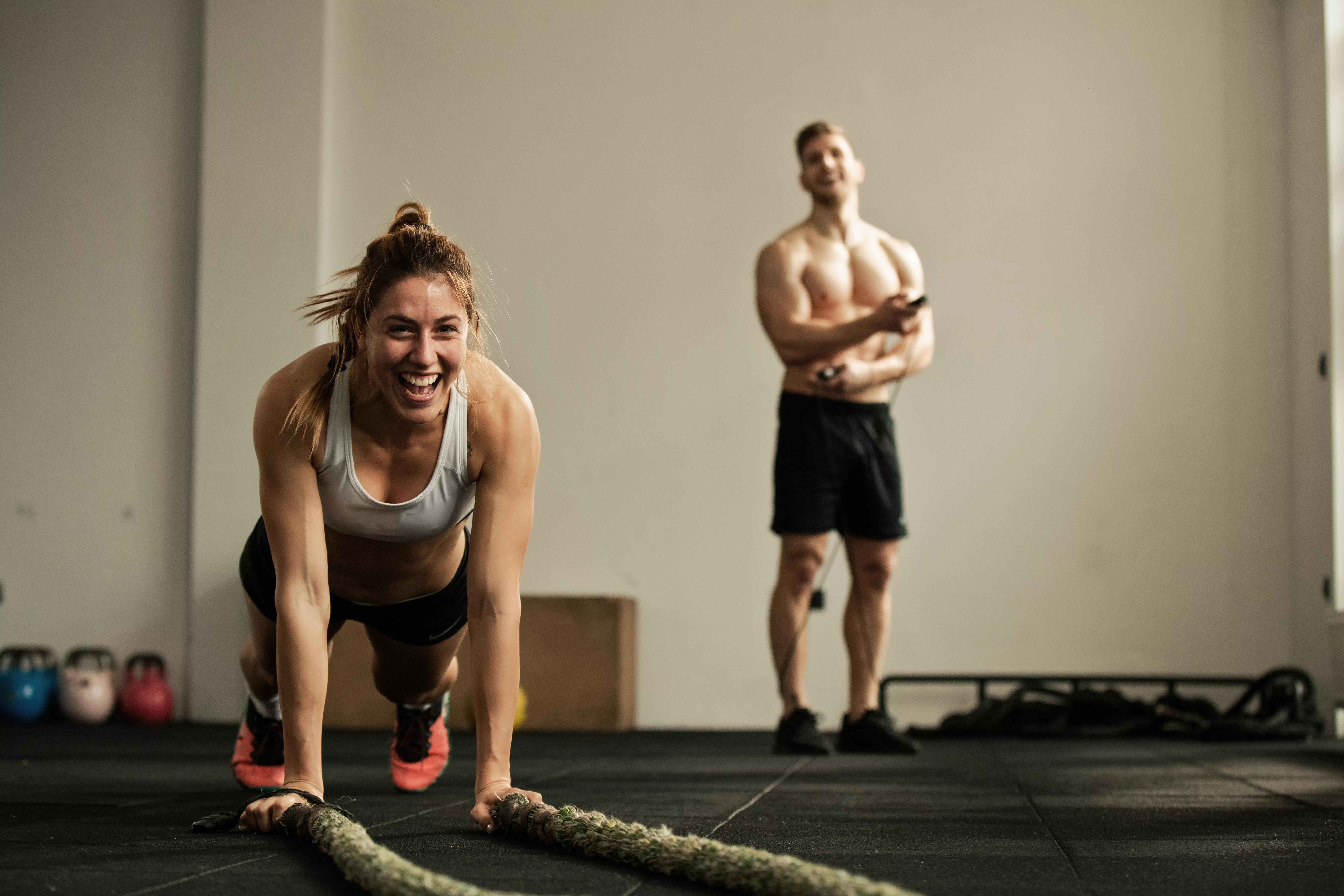 Cheerful female athlete doing push-ups while exercising with battle rope and having fun in a gym.